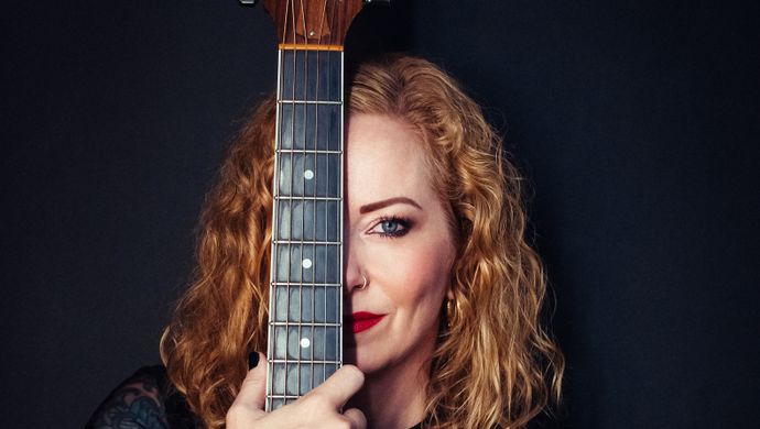 Anneke van Giesbergen kijkt in de camera. Voor haar linker oog houd ze haar gitaar vast. De achtergrond is zwart. 